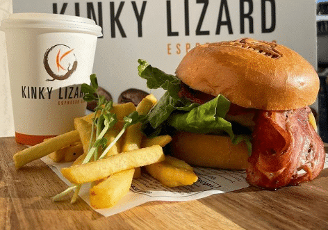 A photo of Kinky Lizard coffee on the left, a bit of fries in the middle and a burger containing bacon and lettuce on the right on a wooden table