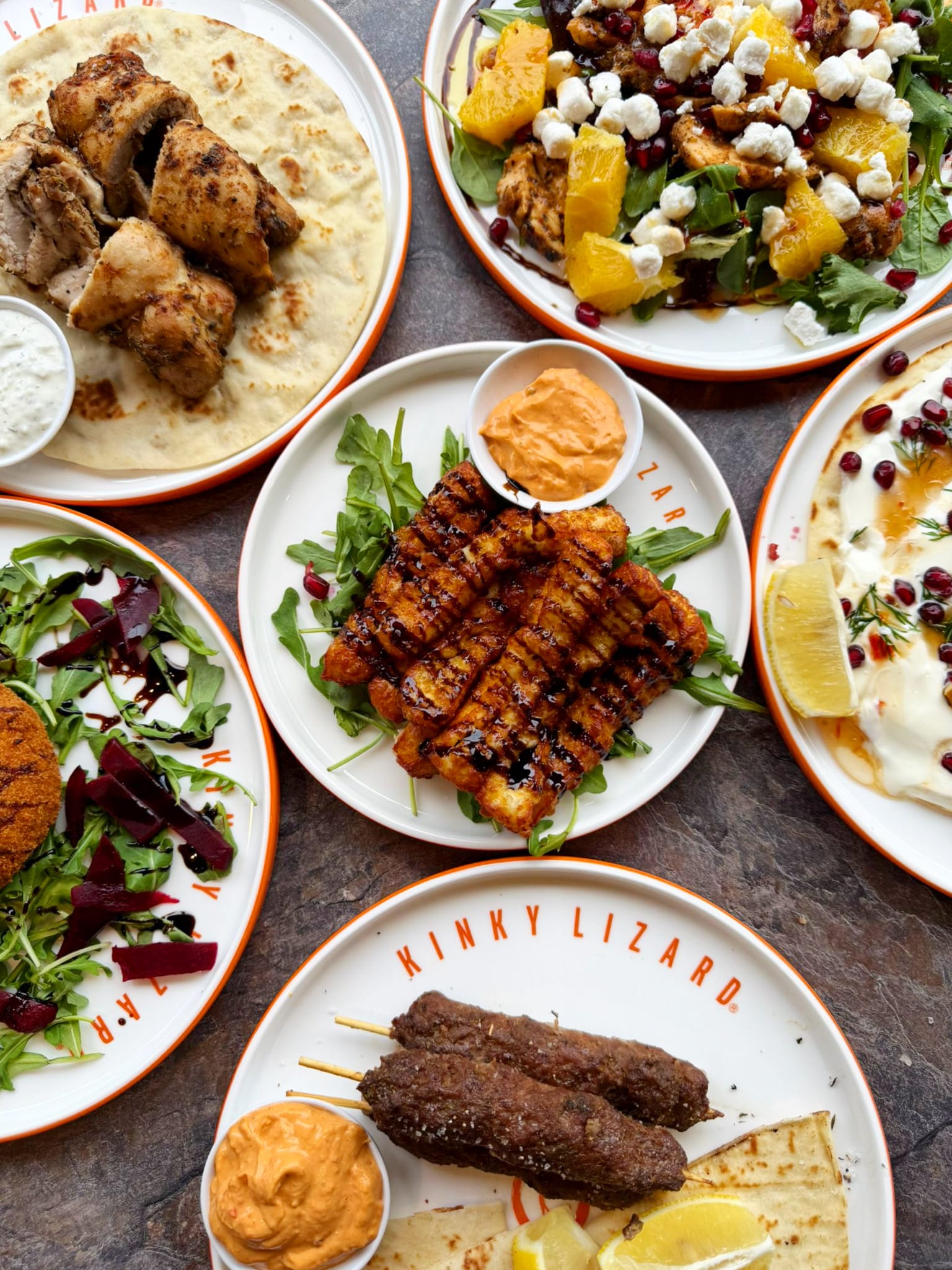 Assorted plates of grilled chicken, flatbreads, and sides with dips and greens on an orange-rimmed table setting; flavorful variety.
