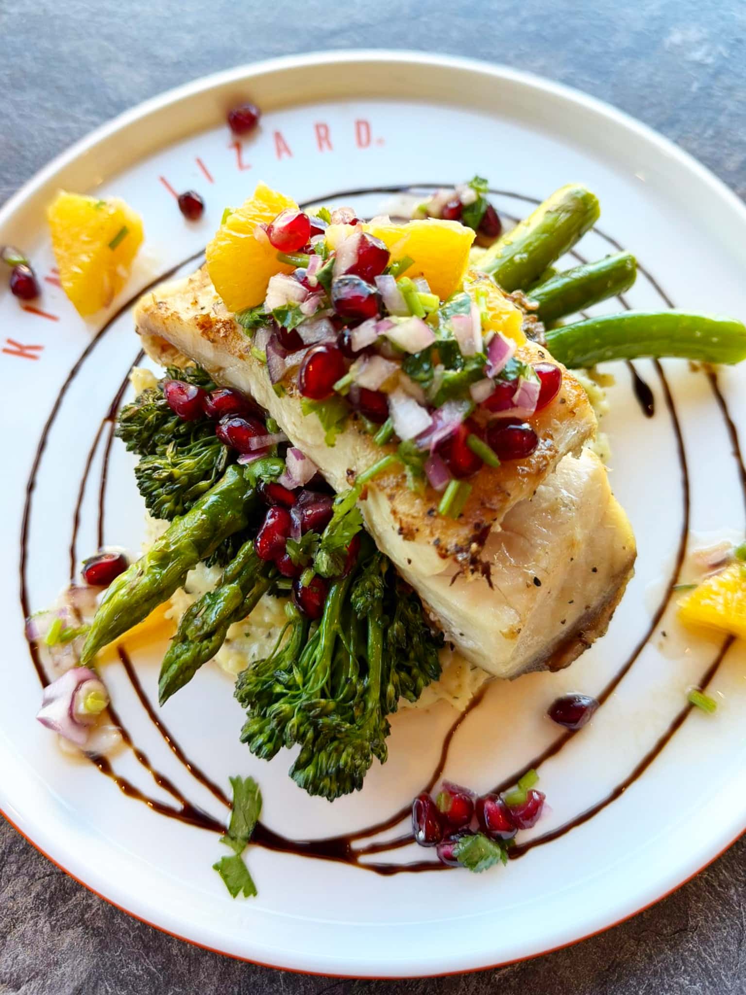 Roasted chicken breast topped with orange segments, pomegranate seeds, red onion, and herbs on a bed of asparagus with a balsamic drizzle.