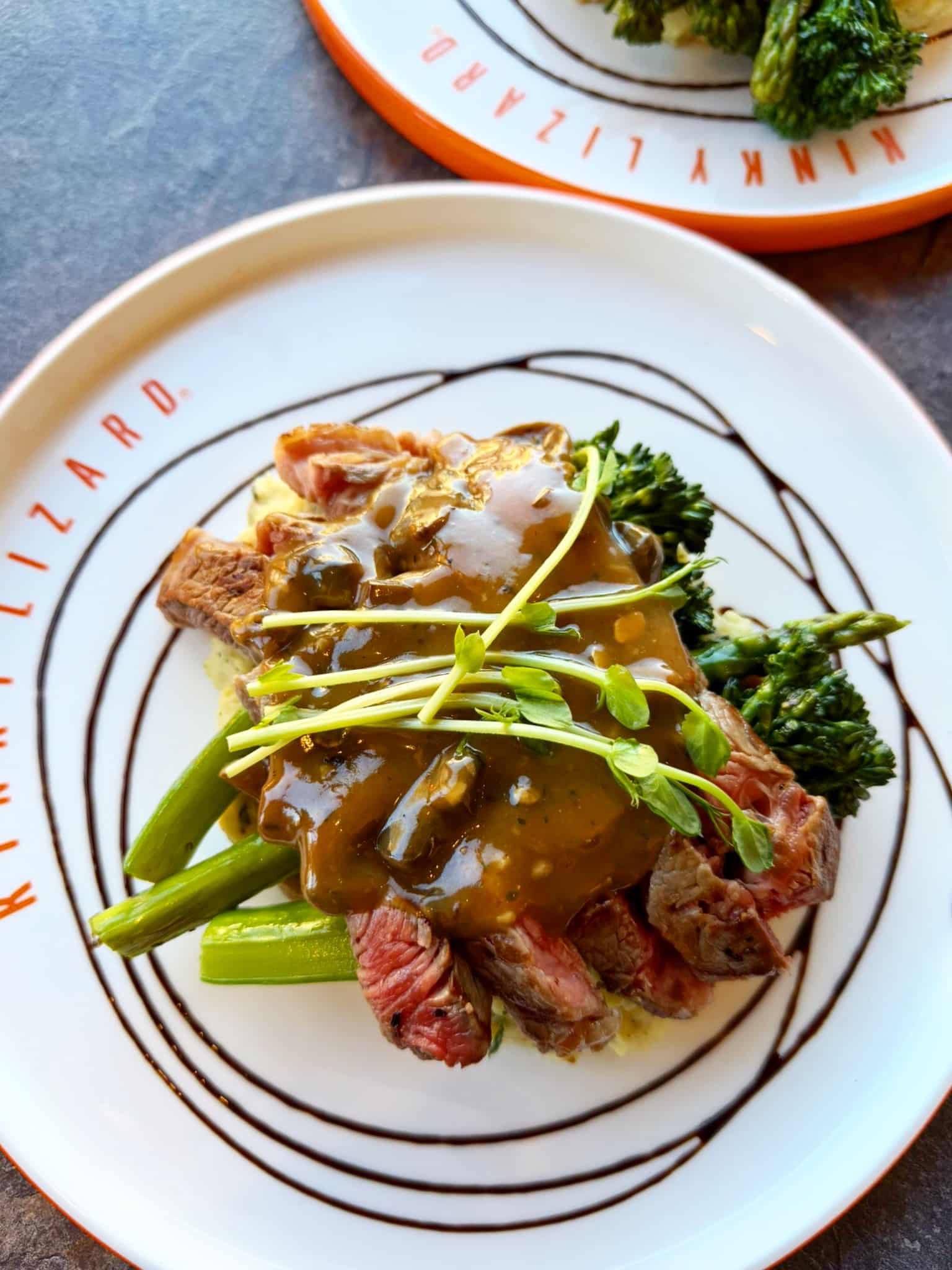 Sliced beef with brown gravy over greens and broccoli, plus asparagus on a decorative plate.