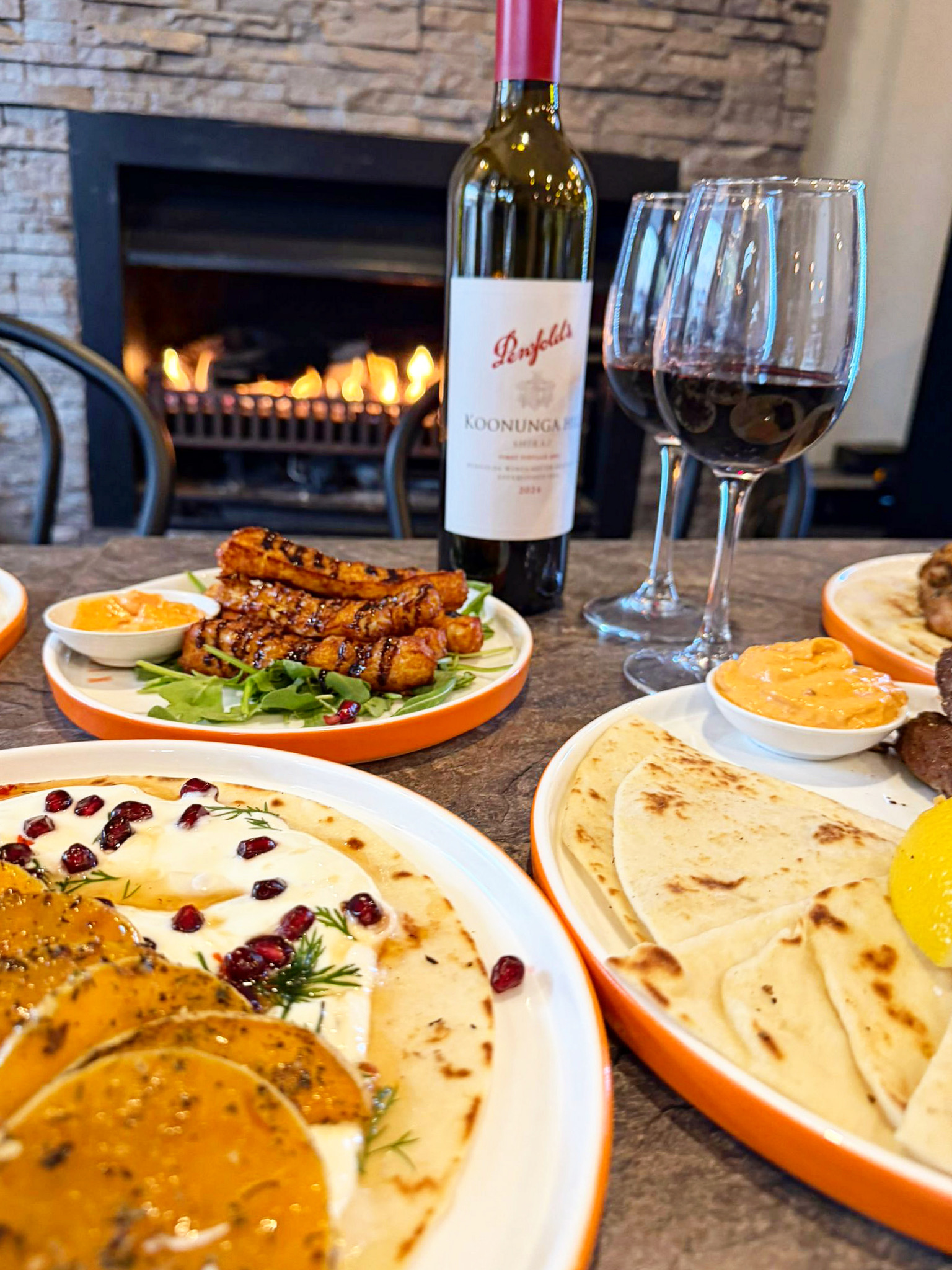 Wine bottle and two glasses with plates of naan, dips, and grilled chicken skewers on a table by a lit fireplace.