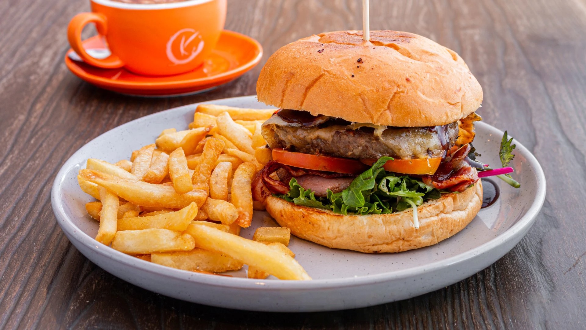 Plate of a stacked burger with lettuce, tomato, bacon and melted cheese, plus a generous serving of french fries on the side, all on a rustic wooden table (orange cup in background).