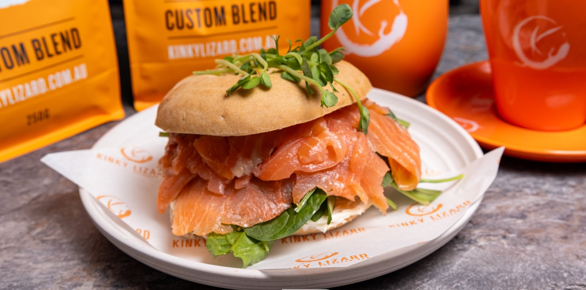 Salmon sandwich on a bun with greens and a sprig of herbs, served on a white plate with orange branding in the background.